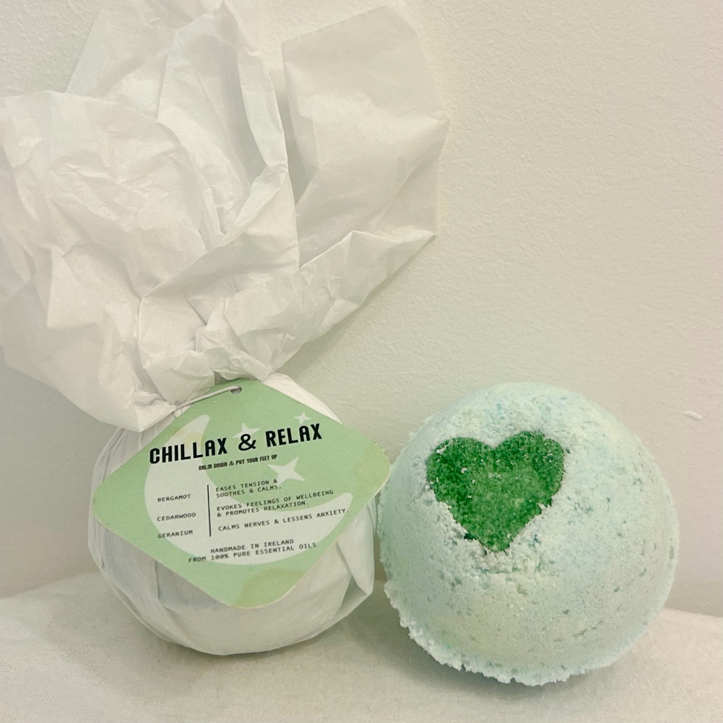 "A round Chillax & Relax Bath Bomb with a white tag displaying the product name; made with 100% natural ingredients and infused with geranium, bergamot, and cedarwood essential oils for a grounding and calming bath."