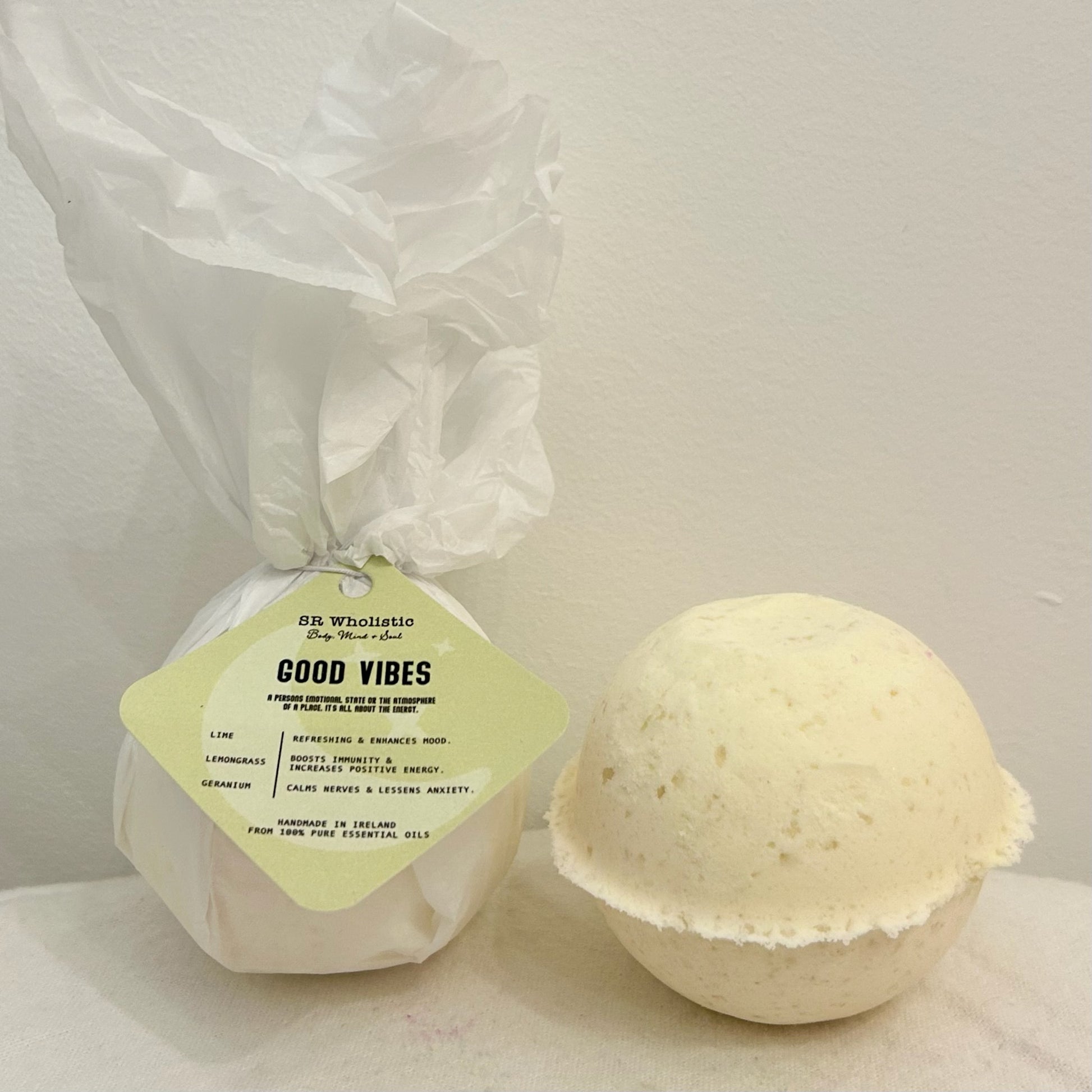 "A round GoodVibes Bath Bomb with a white tag displaying the product name; made with 100% natural ingredients and infused with lemon, lime, and geranium essential oils for an energizing, mood-boosting bath."