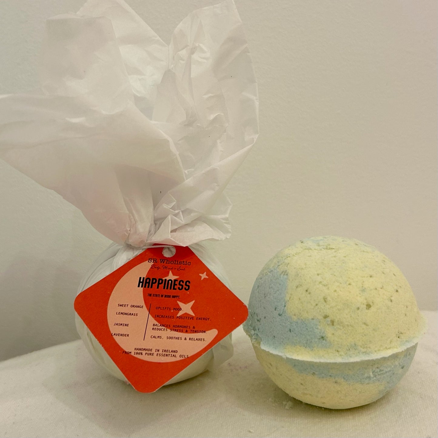 "A round Happiness Bath Bomb with a white tag displaying the product name; made with 100% natural ingredients and infused with jasmine, lavender, orange, and lemongrass essential oils for an uplifting, feel-good soak."