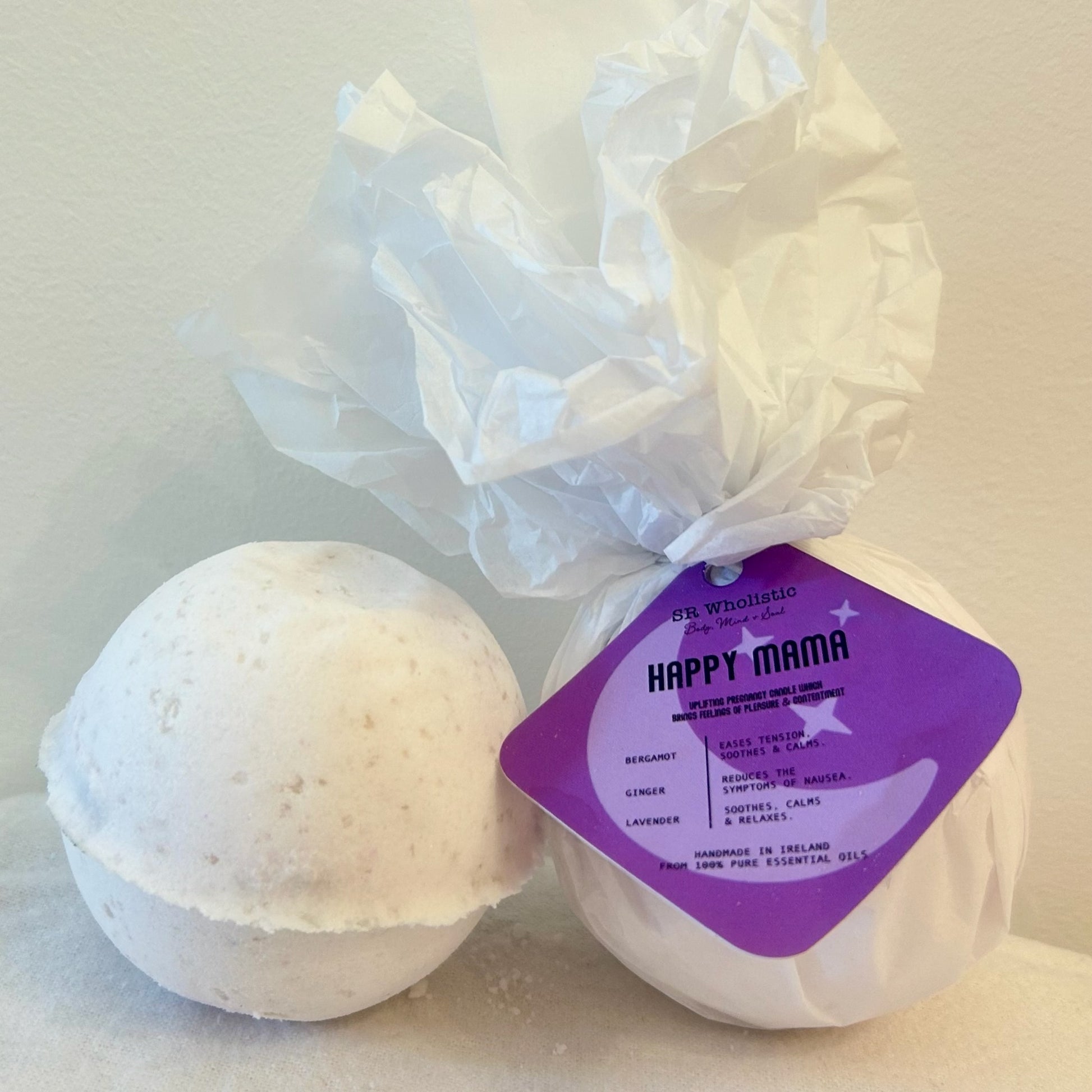 "A round Happy Mama Pregnancy Bath Bomb wrapped with a white tag showing the product name; made with 100% natural ingredients, containing bergamot, lavender, and ginger essential oils."